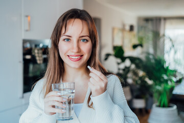 Young smiling woman holding pill and glass of water at home kitchen. Positive lady takes daily...