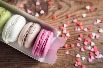 Assorted macarons in a box sprinkled with small hearts on a wooden background, top view