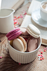 Macaroons in a bowl and a cup of coffee, a book on the background of small hearts on a wooden background, top view