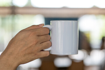 A hand holding out a white blank tea mug with out of focus outdoor cafe background