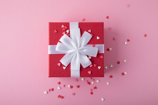Gift box for Valentine's day. Red gift present box with heart confetti on pink background, top view.