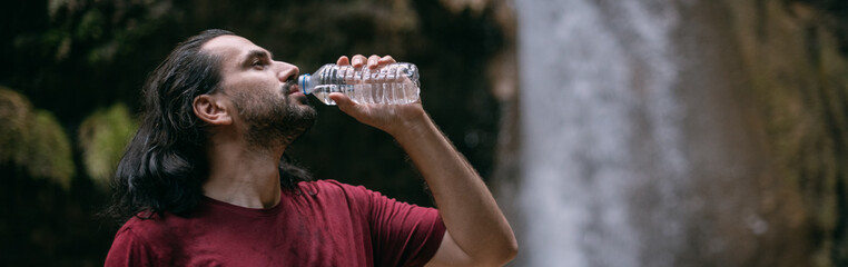 A young man drinks water from a bottle near a tropical waterfall among the rocks.