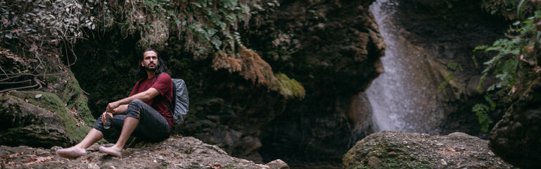 Naklejka na ściany i meble A young male tourist stands near a tropical waterfall among the rocks.