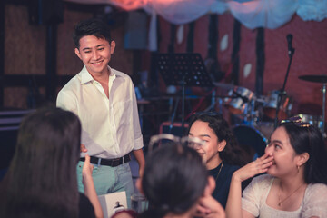 An attractive Asian guy extends his arms to congratulate the lady. A male entrepreneur shakes the hands of the lovely ladies as he welcomes them to his newly opened pub. Restaurant with live band. - obrazy, fototapety, plakaty