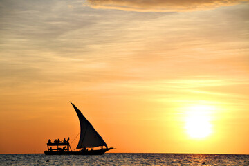 A traditional dhow boat sails through a calm and beautiful blue ocean silhouetted by the setting sun in Zanzibar, Tanzania, Africa.