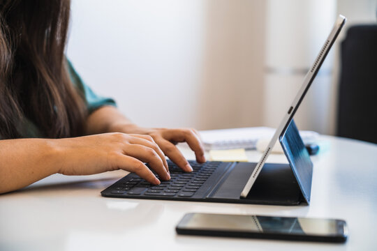 Close up of a woman's hands typing on the keyboard of her hybrid tablet and laptop device next to a phone on a table, while teleworking and adapting to the new normal after coronav