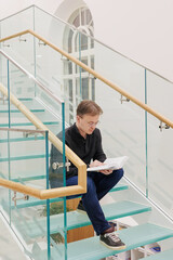 man in library is reading book sitting on steps of glass staircase. education writing scientific work, diploma. thoughtful young adult student or university teacher is learning foreign language