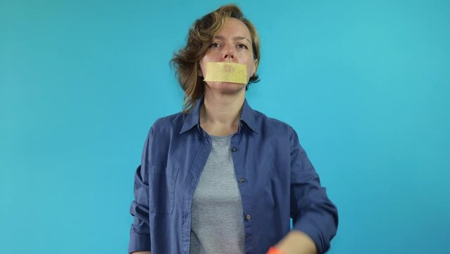 Adult woman in a shirt with a masking tape closed her mouth pointing her finger quietly on an isolated blue background. Woman in orange work gloves. Slow motion portrait