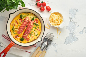 Omelette or frittata with two sausages, melted cheese, cherry tomatoes, broccoli and parsley on red skillet or iron pan on white old concrete background. Healthy breakfast food concept. Top view.