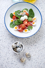 Plate with spinach leaves, cherry tomatoes and quail eggs salad, high angle view on a light-grey granite background, vertical shot