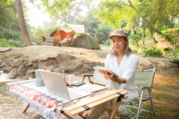 Asian woman travel and camping alone reading a book and using laptop while relaxing on the deck chairs in the river at natural park in Thailand. Recreation and journey outdoor activity lifestyle.