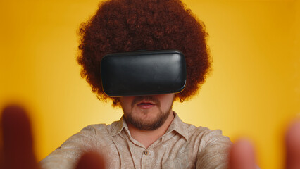 Redhead bearded man with lush afro hairstyle using headset helmet app to play simulation innovation game. Watching virtual reality 3D 360 video. Young guy boy in VR goggles on yellow studio background