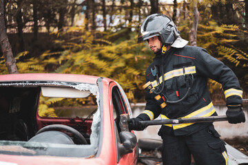 A young and uniformed firefighter breaking the glass of a car that has suffered an accident with a...