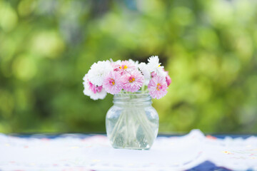 Glass jar with daisies (bellis perennis) on a table in the garden. Congratulations on Easter, greeting card. Copy space. Selective focus.