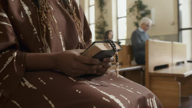 Selective focus medium section shot of unrecognizable Black woman holding prayer book and rosary sitting on bench in church