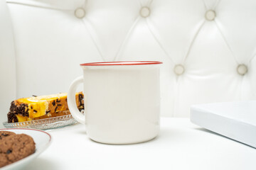 A white blank enamel mug standing out on the top of a white sofa with some snacks placed near it