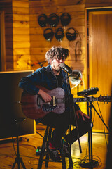 Young rocker musician with messy hair playing a wood electro acoustic guitar and singing in a music studio