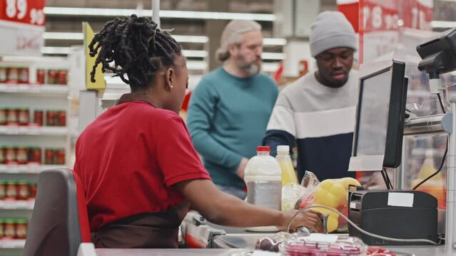 African American female cashier in uniform and queue of customers at checkout in supermarket
