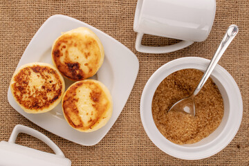 Three homemade cheese pancakes on white ceramic saucer with two cups and sugar bowl, wooden spoon...