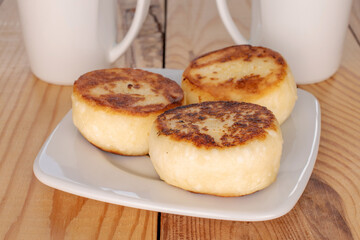 Three homemade cheese pancakes on a white ceramic saucer with white cups on a wooden table, macro.