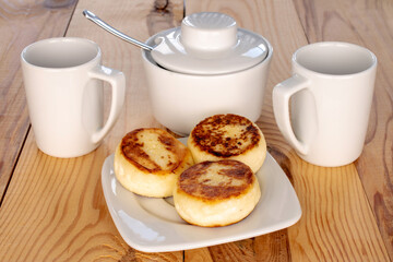 Three homemade cheese pancakes on a white ceramic saucer with white cups on a wooden table, macro.