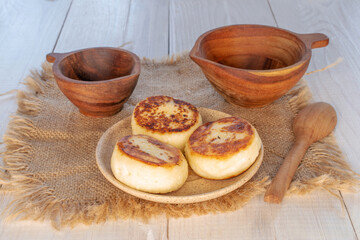 Three homemade cheese pancakes on ceramic saucer with jute napkin and wooden cups on wooden table, macro.