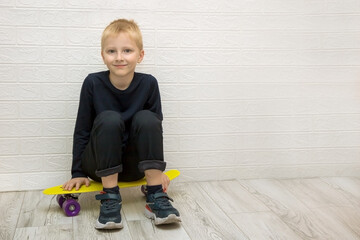tired smiling cute blond boy sitting on a skateboard after training against a white wall