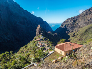 Canary Island, Tenerife view on picturesque Masca village with old stone houses, palm tees, beautiful green sharp hills, cliff, sea horizon and blue sky background