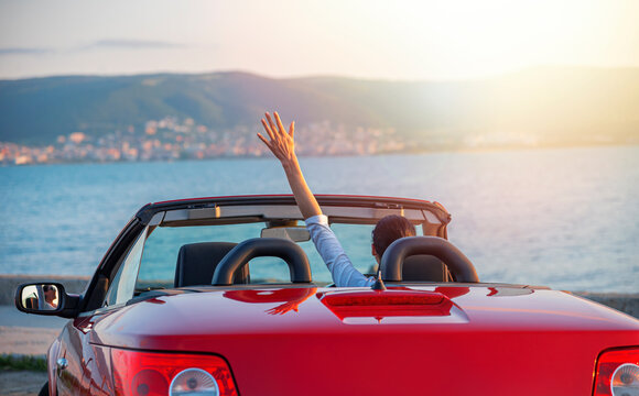 Woman in red car with open roof at background of sea water. Travel, freedom and holidays concept.