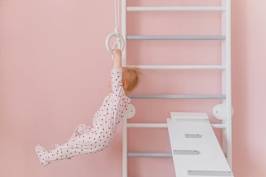 A  toddler girl in soft pink pajamas does gymnastic at home wall bars