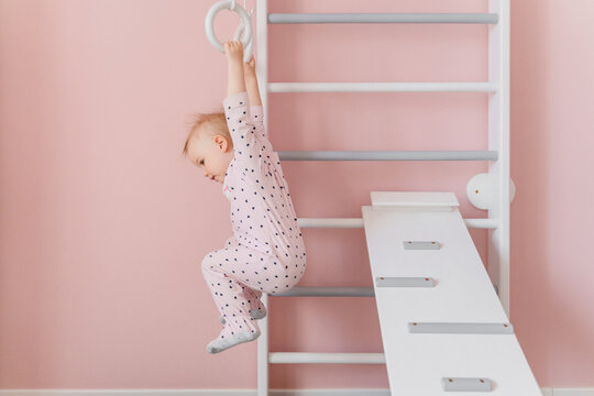 A  toddler girl in soft pink pajamas does gymnastic at home wall bars