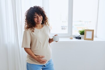Fototapeta na wymiar A pregnant woman with curly hair smiling stands at the window with a mug of warm water and tea and enjoys the view in a home t-shirt, happiness of motherhood