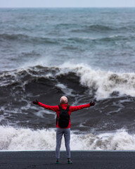 Fearless strong woman with outstretched arms faces powerful waves on black beach; concept of strong...