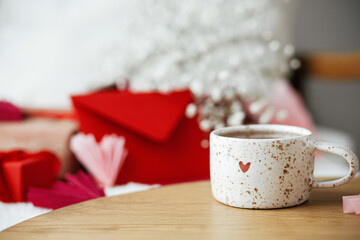 Happy valentines day! Cute modern cup of tea with heart on wooden table against stylish chair with gifts, red envelope and white flowers. Valentine morning surprise for beloved. Space for text