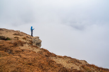 Tourist takes pics on mountain near abyss edge on high altitude under cloudy sky in foggy day. Man...