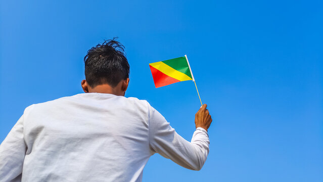 Boy holding Republic of the Congo flag against clear blue sky. Man hand waving Congo-Brazzaville flag view from back, copy space - Powered by Adobe