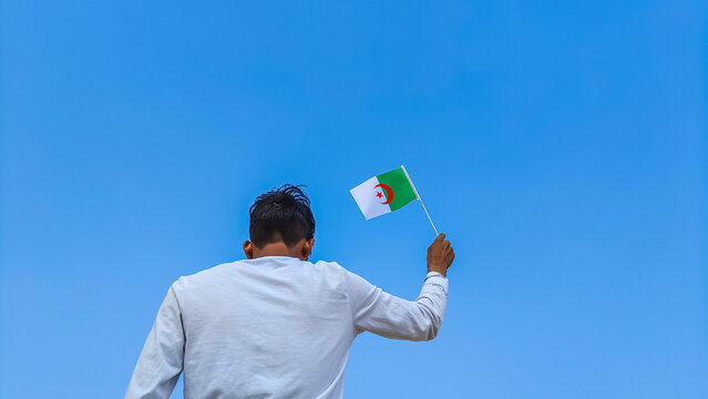 Boy holding Algeria flag against clear blue sky. Man hand waving Algerian flag view from back, copy space