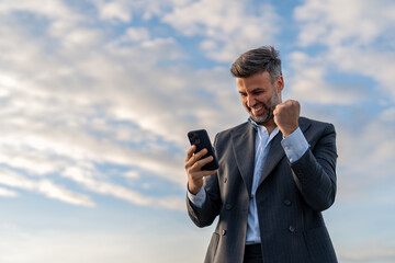 Handsome happy businessman feeling excited raising fist in yes gesture looking at cell phone...