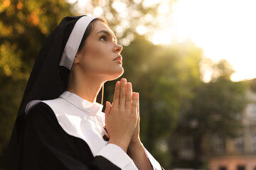 Young nun with hands clasped together praying outdoors on sunny day, space for text