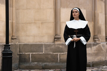 Young nun with Christian cross near building outdoors. Space for text