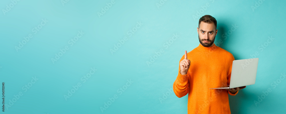 Wall mural Image of adult man teaching online, showing finger while using laptop, explaining rules, standing over light blue background