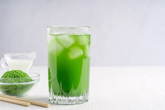 Homemade natural antioxidant matcha made of ground powder of green tea leaves mixed with milk served in glass with ice cubes and paper straw on white wooden table for breakfast. Image with copy space