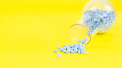 Blue pills falling out of a glass flask on yellow background Space for text on the left