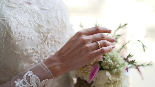 Elegant bride in fashion lace wedding dress holding wedding bouquet of fresh flowers and touching with her tender hand, classic manicure and gold wedding ring on bride's finger, emotional details of