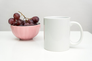 A white blank coffee mug on the top of a white table with the grapes in a bowl placed behind it