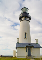 A white lighthouse and a small house under a cloudy sky on a green lawn. Majestic architecture, navigation, history, cultural heritage, travel, tourism.