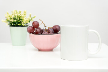 A white blank coffee mug on the top of a white table with the grapes in a bowl placed behind it