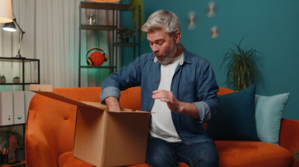 Happy surprised winner man unpacking delivery parcel at home. Smiling satisfied mature guy shopper...