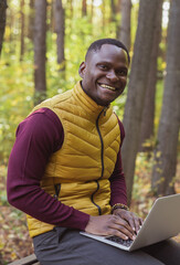 Portrait African american man sitting in city park on a bench with laptop studying online outdoors. Man freelancer learn working remotely in street e-learning vertical banner