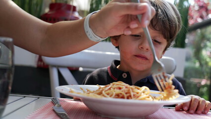 Child sitting at lunch table eating spaghetti. Mother hand feeding pasta to son at restaurant. Noodles with red sauce on plate